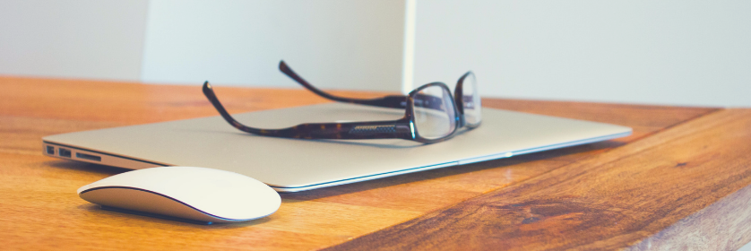 A closed Macbook, Apple mouse, and a pair of glasses.